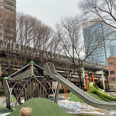 Climbing structure, two slides, hills, and a passing L-train on the green or orange line. The orange posts are the saucer swing.