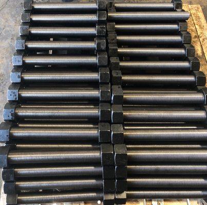A large batch of precision-machined heavy-duty stud bolts with hex nuts, arranged in uniform rows...