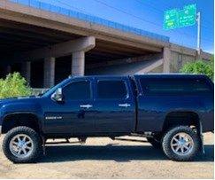 A blue Silverado truck with a camper shell parked in front of an overpass, highlighting its lifted stance and bold look.