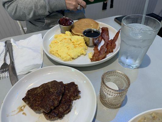 Brunch Pancakes with strawberry compote, maple syrup, bacon, and eggs scrambled eggs. Side of house made sausage patties in foreground