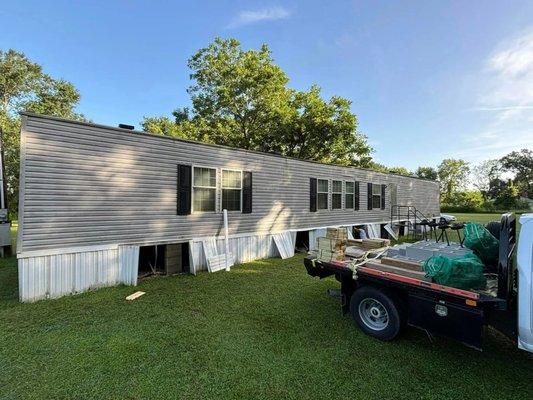A wide shot of a mobile home with partially installed skirting, with a flatbed truck loaded with materials in the foreground.