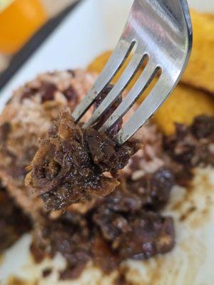 Close-up of a fork holding a piece of my Oxtail, with Rice and Peas and Sweet Plantains in the background.
