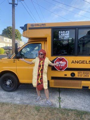A person dressed as a hot dog standing in front of a camp bus