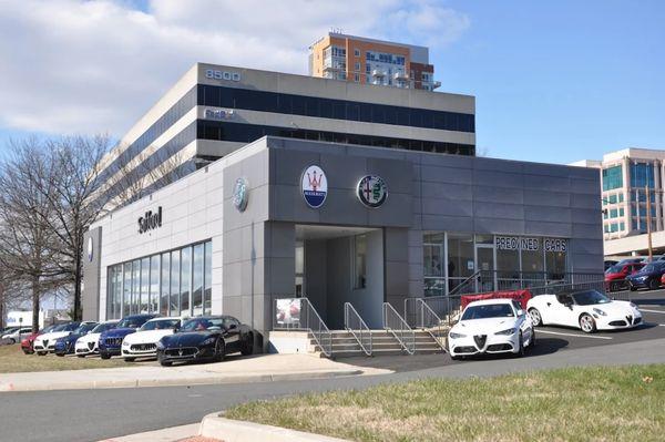 Exterior of Alfa Romeo Tysons Corner and Maserati dealership in Tysons VA, featuring glass showroom, preowned cars, and city office towers.