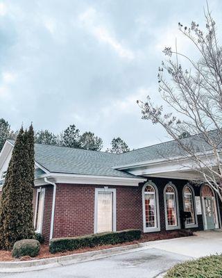 A brick building with a gray shingle roof and arched windows is seen under a cloudy sky, with landscaping and bare trees.