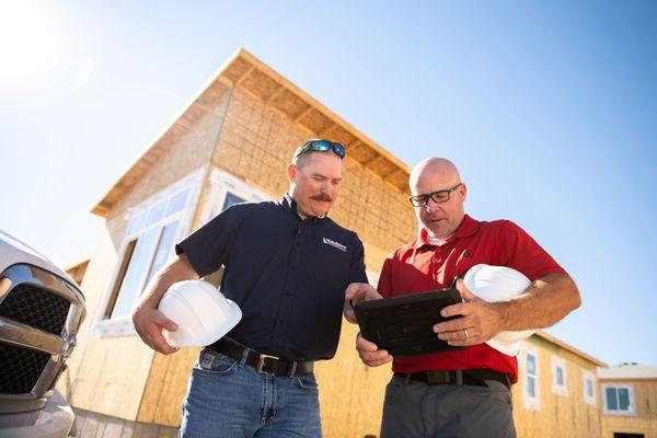 Two Builders FirstSource team members hold hard hats while reviewing construction plans on a tablet...