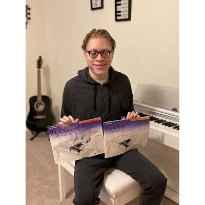 This photo features a smiling student proudly showcasing two Piano Adventures lesson books while seated at a sleek white digi...