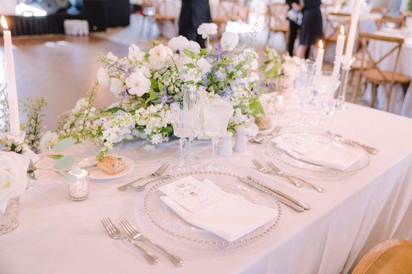 An elegant and romantic tablescape featuring delicate floral arrangements in soft whites and hints of lavender. Crystal-clear charger plates