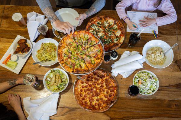 A table with three pizzas, salads, pasta, wings and drinks. Two people are dining, one serving pizza, the other coloring.