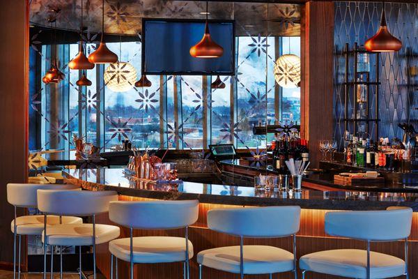 Chic indoor bar at VASO with modern pendant lighting, backlit shelves, and floral-patterned glass framing the city beyond.