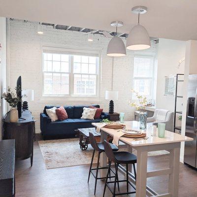 Living room & kitchen in model apartment with high ceilings, movable kitchen island, quartz countertops, windowed cabinets,& large windows.