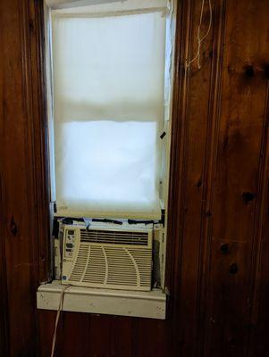 Air conditioner, complete with duct tape, air gaps, and a non-working shade, looking out on trash piled next to an old shed.