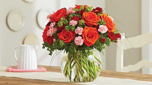A vibrant bouquet of orange roses, red carnations, pink mini carnations, and lush greenery beautifully arranged in a round gl...