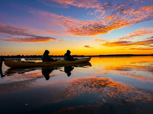A tandem kayak floating on Lake Huron at sunrise with two people and pink clouds reflecting in the calm water.