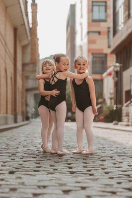 Three young dancers wearing black leotards and pink tights and ballet slippers smile at the camera with their arms around each other