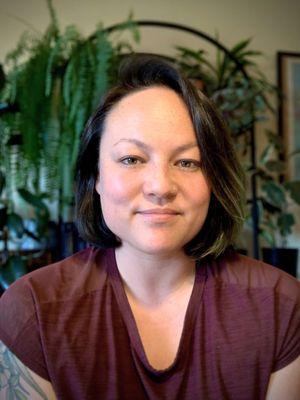 Photo of a biracial ciswoman with medium length dark brown hair and a maroon shirt sitting in front of a bookshelf with several plants.