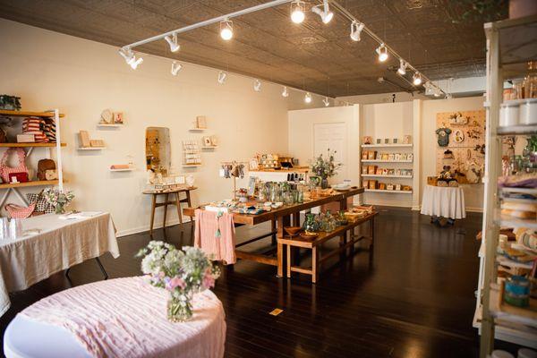 Interior of gift store showing a table with glassware, pottery, candles and more! Jewelry on the wall; cards and coffee on back shelves.