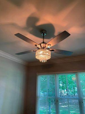 A modern ceiling fan with a crystal chandelier hangs from a white ceiling. The fan has four blades and the chandelier is lit,...