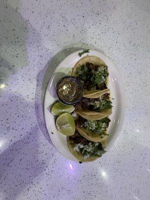 In this photo there are 3 tacos de lengua,with lemons, It also includes cilantro and onion one of my favorite Mexican dishes must try.