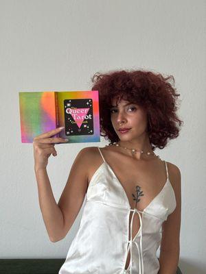 The owner Jery Ruiz posing with their Queer Tarot Deck book, The Curl Parlor offers book-read tarot readings.