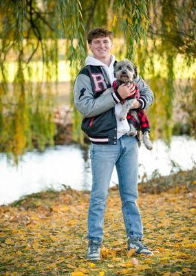 Senior boy standing outdoors by the creek with his letterman's jacket on holding his dog in his hands while standing on fallen leaves.
