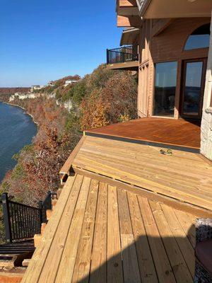 A multi-level cliffside deck is partially finished, showing a stark contrast between the bright, newly installed natural wood...