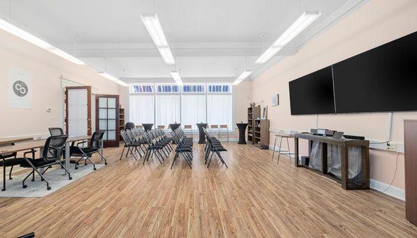 A bright meeting or event room with light wooden floors, pink walls, rows of black folding chairs, and two large, black scree...