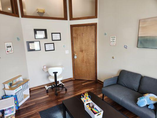 Therapy office with children's toys, art on the wall, certificates, coffee table, blue rug, and couch.