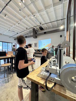 Children of different ages collaborating at workstations with specialized equipment in a bright, industrial-style learning environment