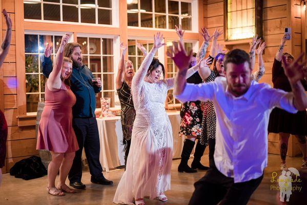 A joyful wedding reception scene. The bride, wearing a white gown, is at the center of the dance floor with her arms raised.