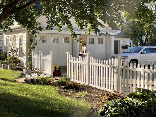 A beautiful photo of the front of a house. The side yard is lined with a white picket fence and a large tree is casting a shadow.