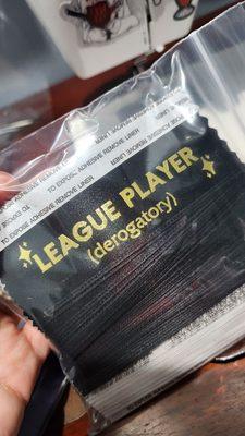 bag of black badge ribbons with gold print that says "LEAGUE PLAYER (derogatory)" with sparkle stars around it.