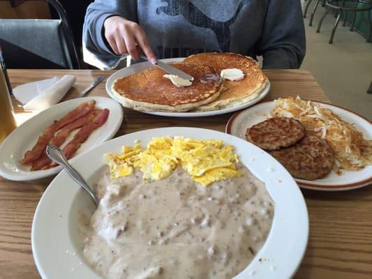 Biscuit covered in sausage gravy and scrambled eggs. Side of bacon, side of sausage and hash browns. Buttermilk pancakes in background.