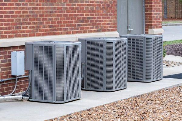 Three large outdoor air conditioning units installed beside a brick building, connected to electrical wiring and refrigerant ...