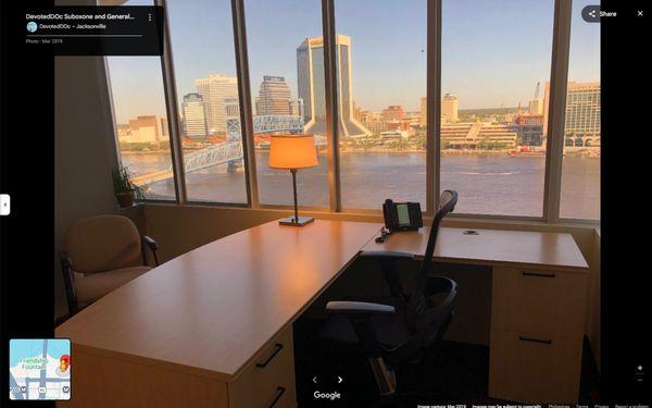 Private office space with a scenic city and river view, featuring a desk, chairs, and warm natural lighting.