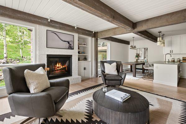 Elegant living room featuring painted white brick fireplace, exposed weathered beams, and seamless transition to dining and kitchen areas. T