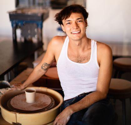 Dax sitting at the pottery wheel with a small cup he made. This is a demonstration of what a beginning class will make to take home .