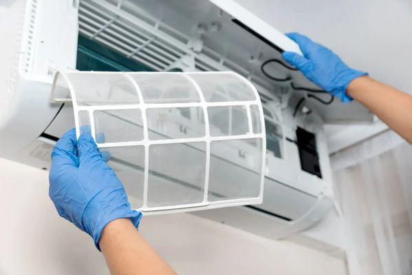 A person wearing blue gloves is removing or replacing a white mesh air filter from the front panel of a white, wall-mounted m...