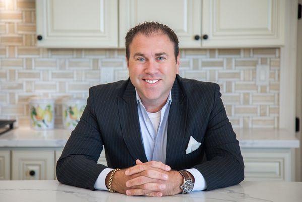 A professional headshot of Charles Hoelzel sitting at a kitchen counter.