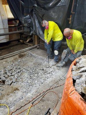 Two workers in safety gear, including masks and knee pads, are using jackhammers to break up a concrete floor.
