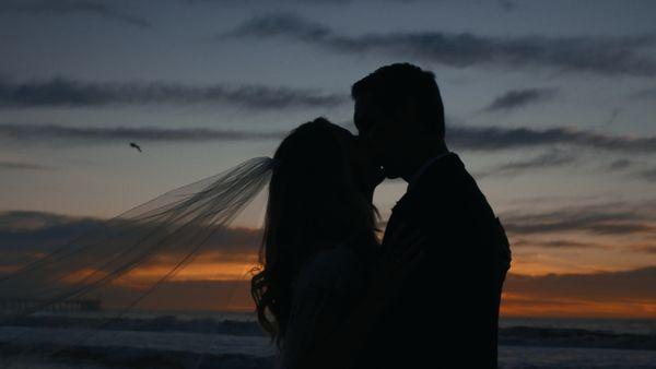 Bride and groom kissing at sunset on the California coast, cinematic wedding videography style with flowing veil and romantic ocean backdrop