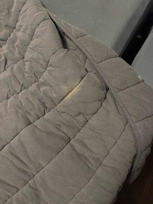 Yellow blotches throughout gray bed covering to also include an imprinted press marking. Additional photo will show small holes.