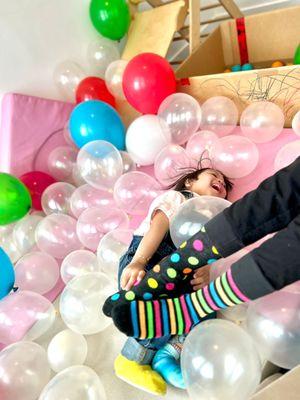 Fotografía de un niño pequeño acostado en el suelo rodeado de un lecho de globos y pies de adultos con calcetines de lunares