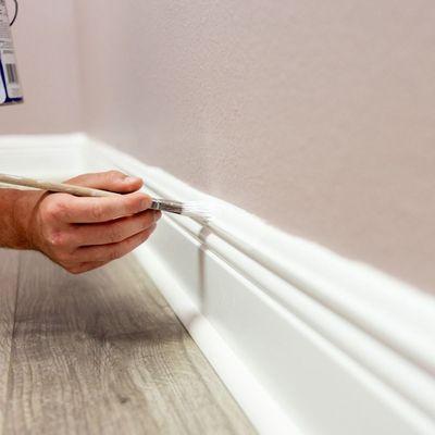 A close-up shows a hand painting a white baseboard with a brush, against a light purple wall and gray wood-look flooring.