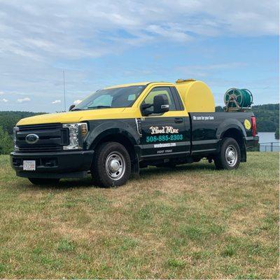 A Weed Man Spencer truck on a grassy hill by a lake. The yellow and green Ford pickup shows their logo, number (508-885-2303).