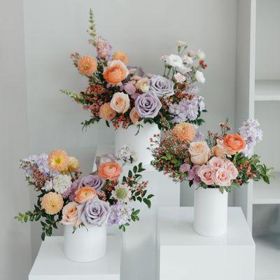 Chic and modern trio of floral arrangements featuring soft lavender roses, peach blooms in white ceramic vases. Perfect for weddings!