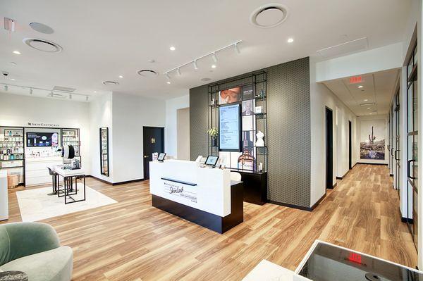 A modern, open-concept space featuring our SkinCeuticals retail wall, check-in desk, and hallway leading to private treatment rooms.