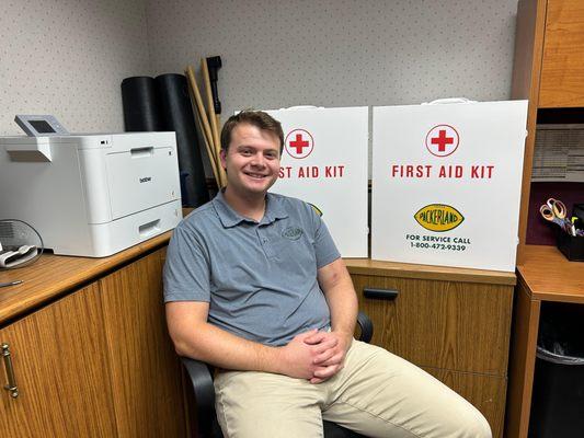 A Packerland employee wearing a grey collared shirt and khaki pants sits on an office chair with his hands casually folded on...