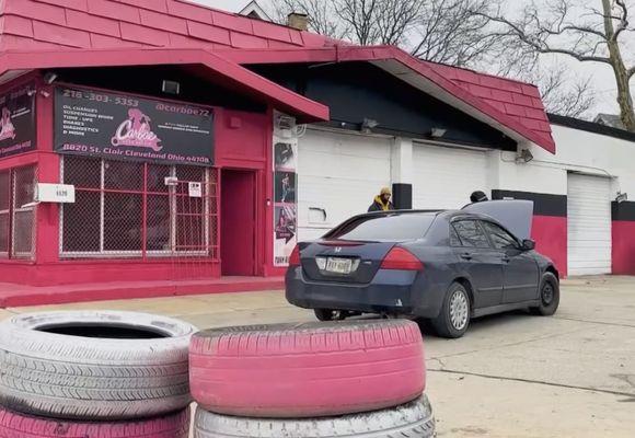 A car is parked in front of a pink-painted auto mechanic building. The car's hood is open, and a mechanic is looking inside with a bystander