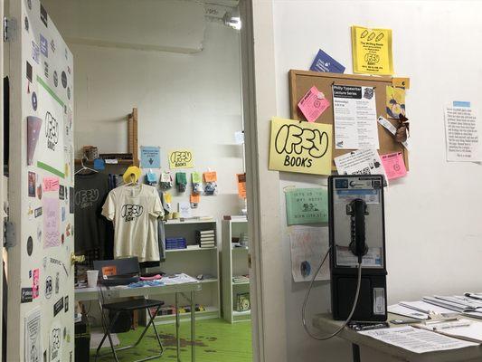 The entrance to Iffy Books, with a door covered in stickers. There are books, t-shirts, and electronics inside. A payphone sits on a table.